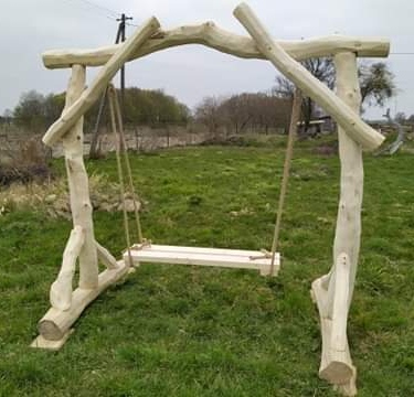 Drewniana huśtawka ogrodowa wykonana z naturalnych gałęzi, umieszczona na trawniku, widoczny słup energetyczny w tle.