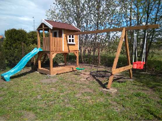 Drewniany plac zabaw z domkiem na podeście, zjeżdżalnią, huśtawką typu bocianie gniazdo i klasyczną huśtawką z czerwonym siedziskiem, ustawiony na trawniku w ogrodzie.
