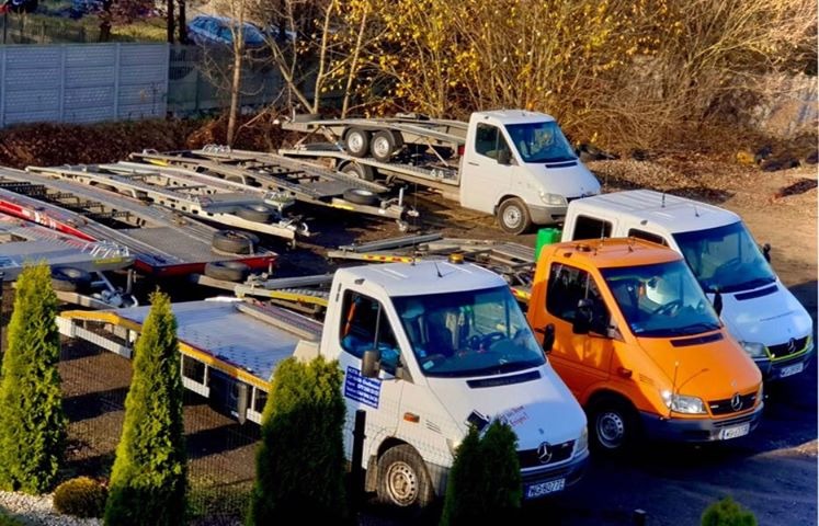 Flota pojazdów: białe i pomarańczowe busy z przyczepami do transportu aut, zaparkowane na placu w jesiennym słońcu.