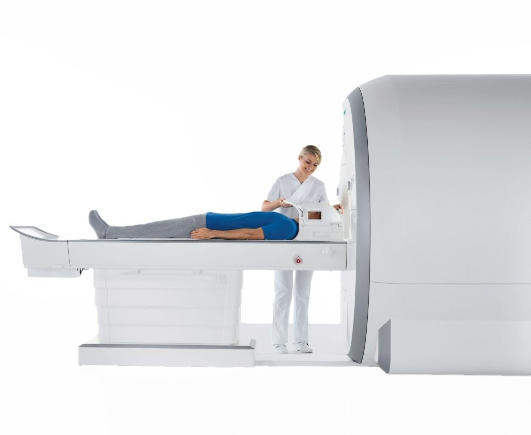 Pacjent w skarpetach i niebieskiej koszulce leży na stole tomografu, obok stoi uśmiechnięta pielęgniarka w białym uniformie przygotowująca urządzenie.