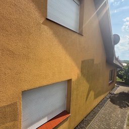 Dawid Kojka - Fragment żółtej elewacji budynku z widocznym efektem mycia, gdzie ciemniejszy obszar kontrastuje z resztą. Dwa okna z białymi roletami i czerwonym parapetem.