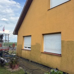 Dawid Kojka - Żółta elewacja domu z widoczną linią podziału między czystą a zabrudzoną częścią, obok okien z roletami. Na trawniku sprzęt do czyszczenia, w tle podnośnik z polską flagą.