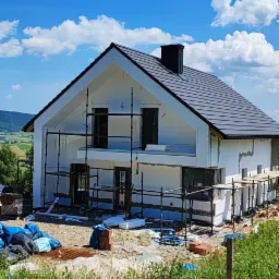 Dom jednorodzinny w trakcie ocieplania, widoczne rusztowania, niezabezpieczone materiały budowlane, w tle zielony krajobraz.