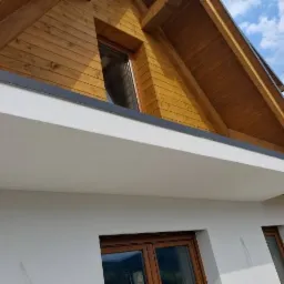 Fragment domu z drewnianą konstrukcją dachu i białą elewacją z widocznymi oknami w drewnianych ramach.