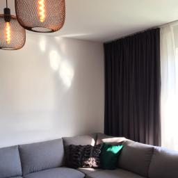 Malarz Mieszkaniowy - Szary narożnik z poduszkami w salonie, oświetlony trzema lampami z żarówkami Edisona, obok okno z ciemnymi zasłonami i białymi firankami.