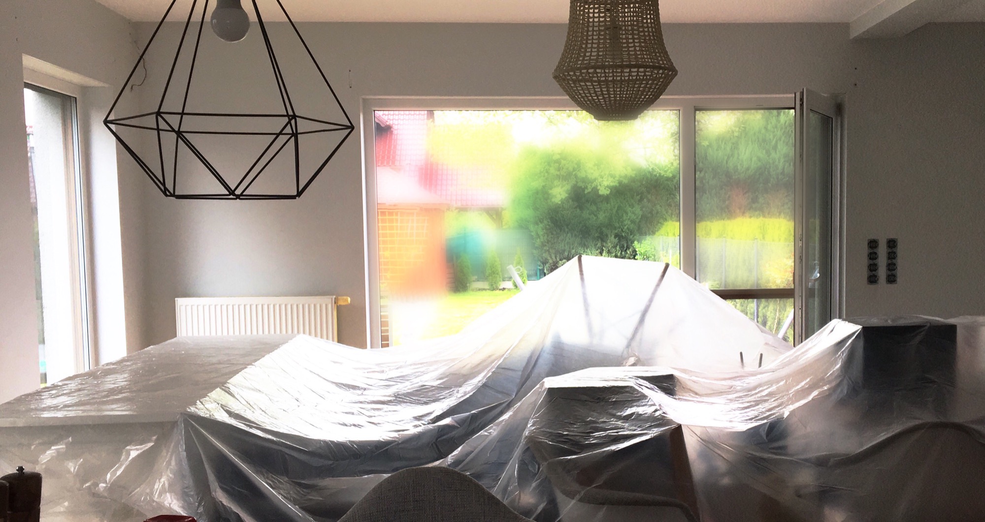 Pokój zabezpieczony folią malarską przed malowaniem, widoczne dwa typy lamp, geometryczna i rattanowa, okno z widokiem na ogród.
