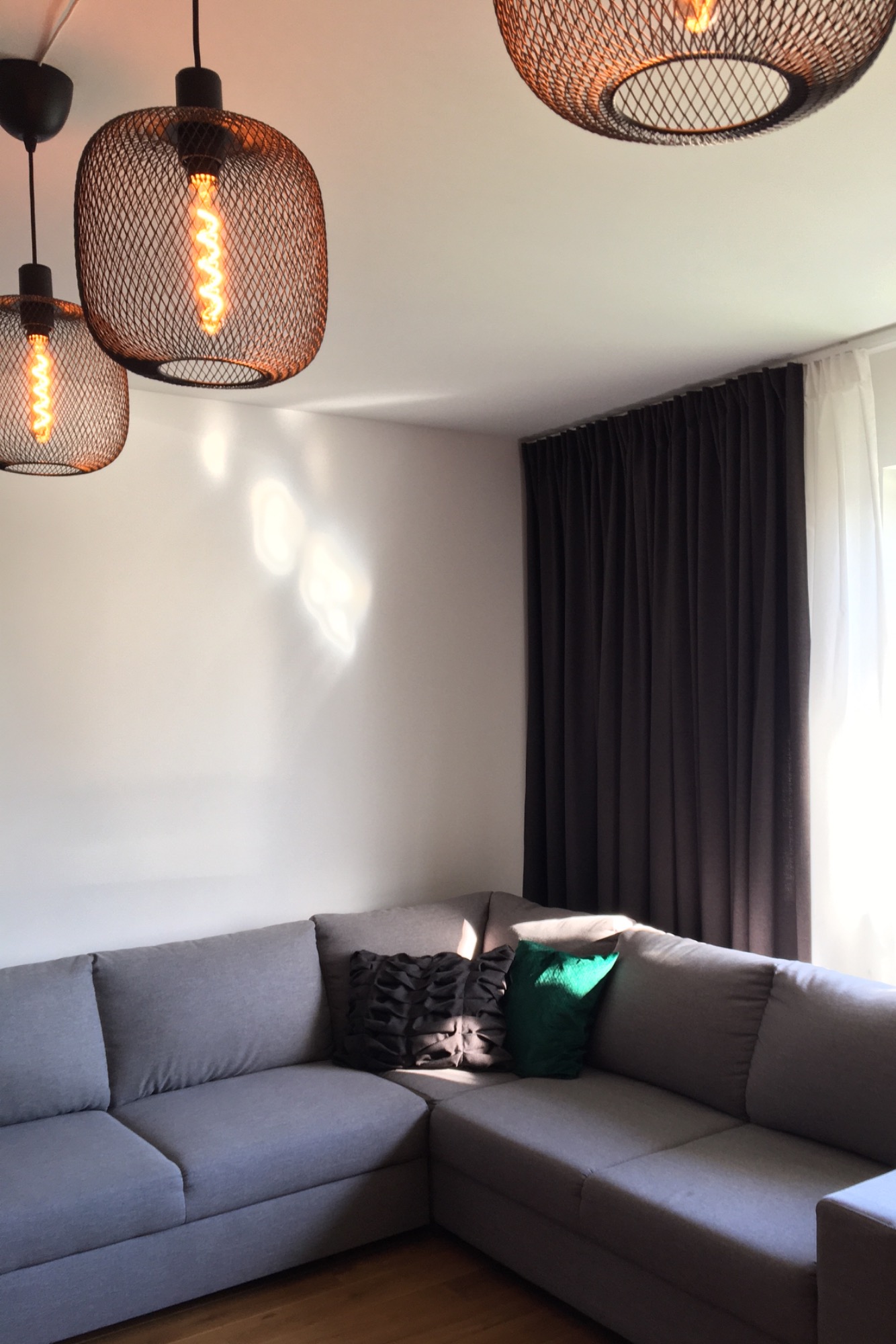 Szary narożnik z poduszkami w salonie, oświetlony trzema lampami z żarówkami Edisona, obok okno z ciemnymi zasłonami i białymi firankami.