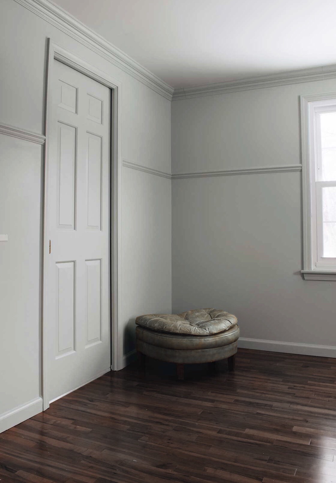 Pokój po odświeżeniu: ściany pomalowane na szaro z listwami dekoracyjnymi, ciemna podłoga z paneli, białe drzwi i okno, skórzany puf na środku.