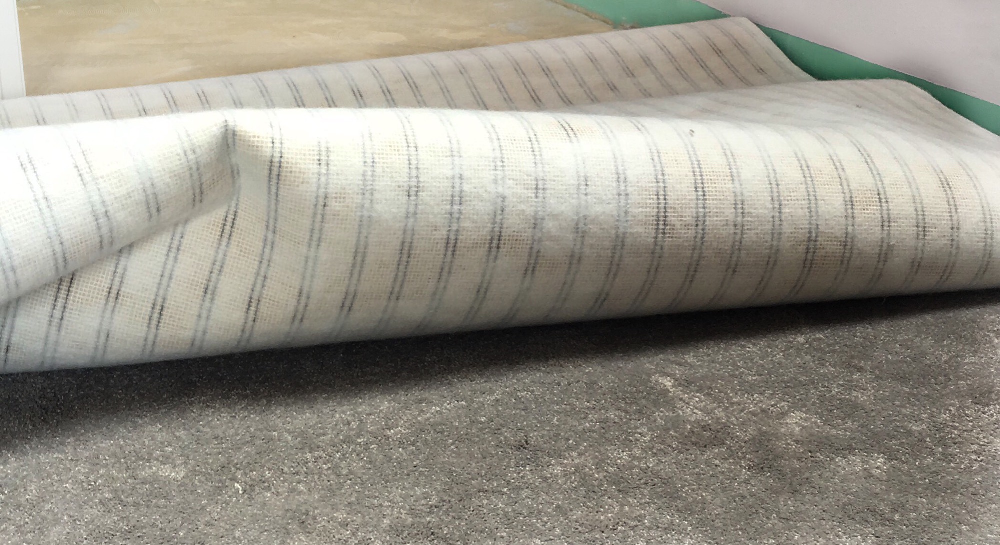 Fragment zwiniętej wykładziny w biało-szare paski leżącej na szarej podłodze, widoczny fragment ściany w tle.