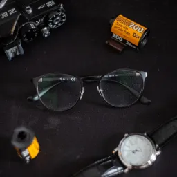 Czarny stół z analogowym aparatem Zenit, zwiniętą kliszą 35mm, okularami Ray-Ban i zegarkiem na skórzanym pasku.