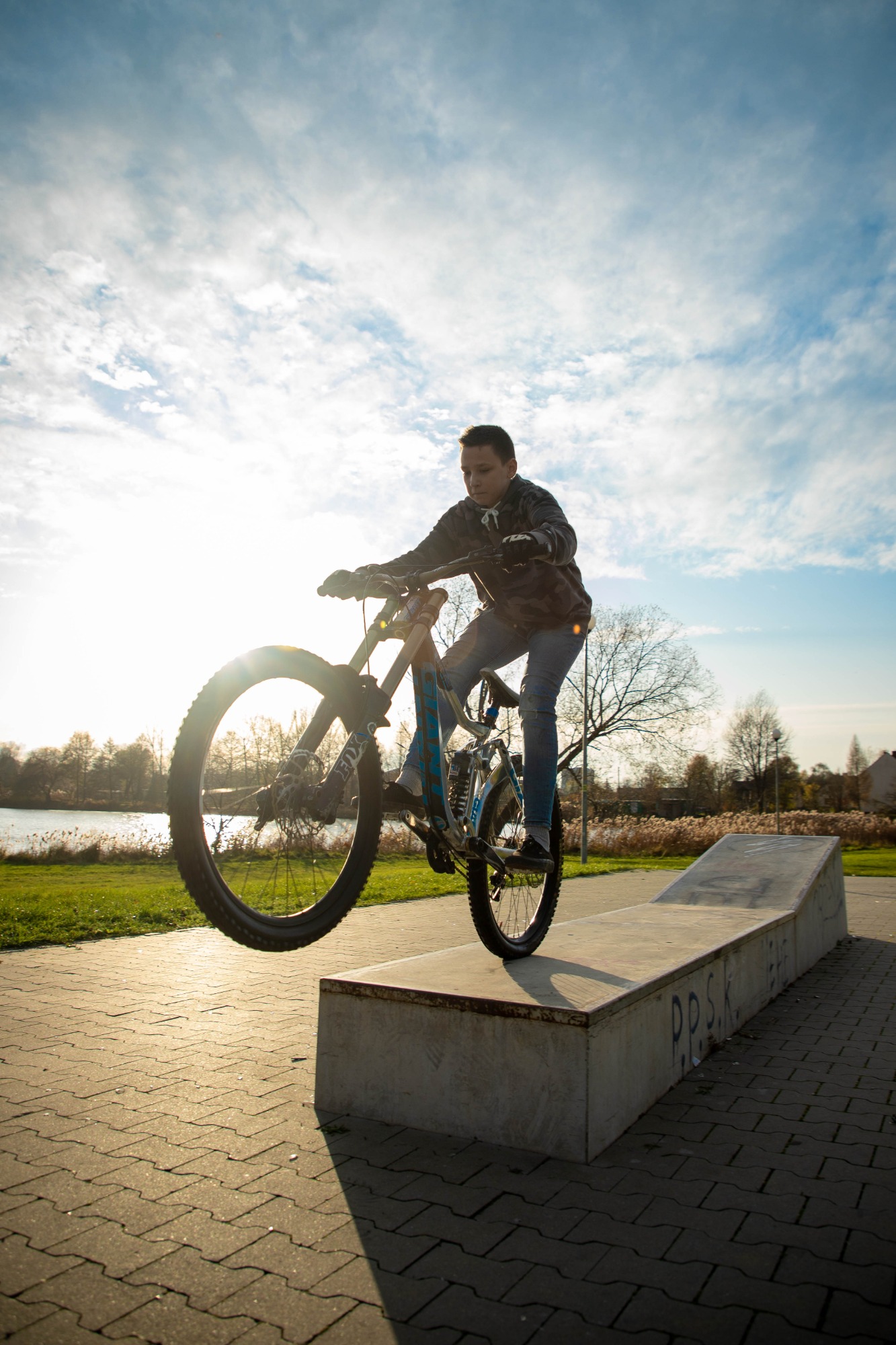 Młody rowerzysta wykonuje trik na rampie w skateparku, rower górski z amortyzatorami, w tle jezioro i zachodzące słońce.