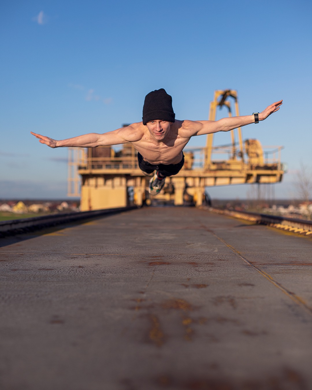 Mężczyzna w czapce, bez koszulki, w locie na tle metalowej konstrukcji i błękitnego nieba.