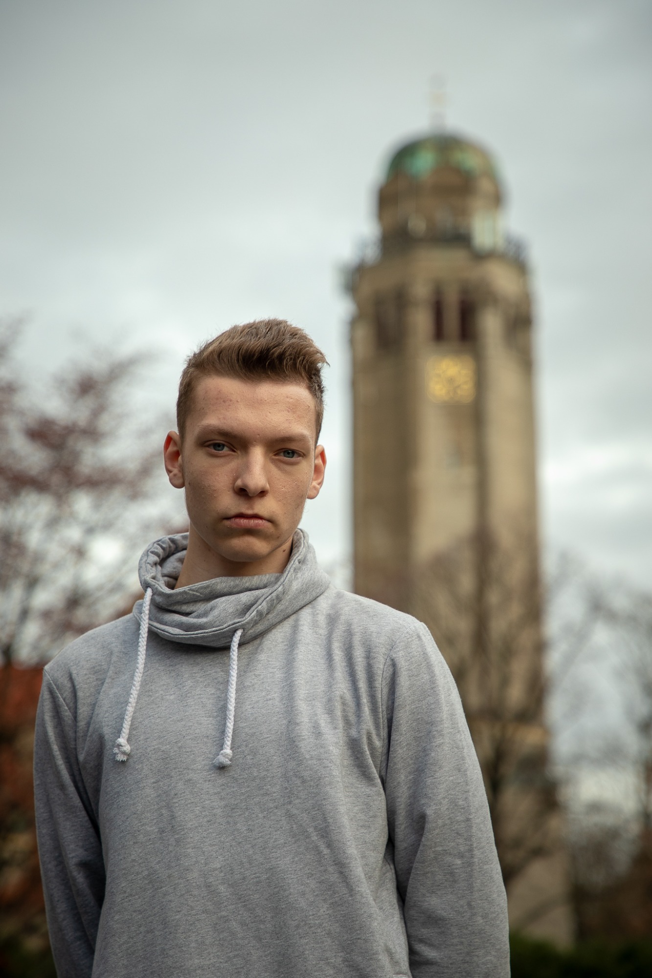 Portret młodego mężczyzny w szarej bluzie z kapturem na tle rozmytej wieży zegarowej z zieloną kopułą.