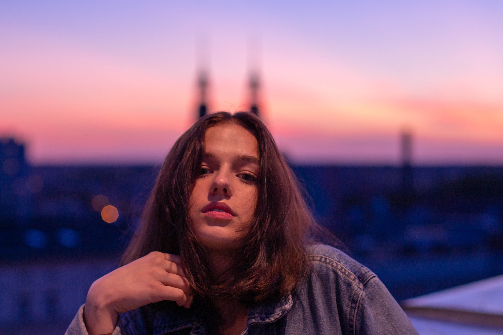Portret młodej kobiety w jeansowej kurtce na tle zachodzącego słońca zarysowującego sylwetki budynków
