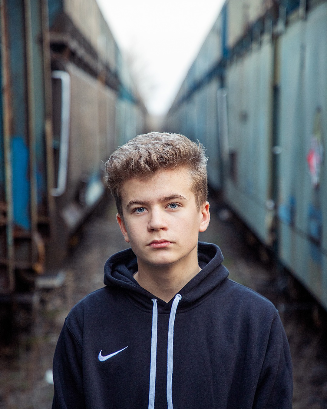Portret młodego mężczyzny z niebieskimi oczami, ubrany w czarną bluzę z kapturem, stojącego między dwoma wagonami kolejowymi na torach.
