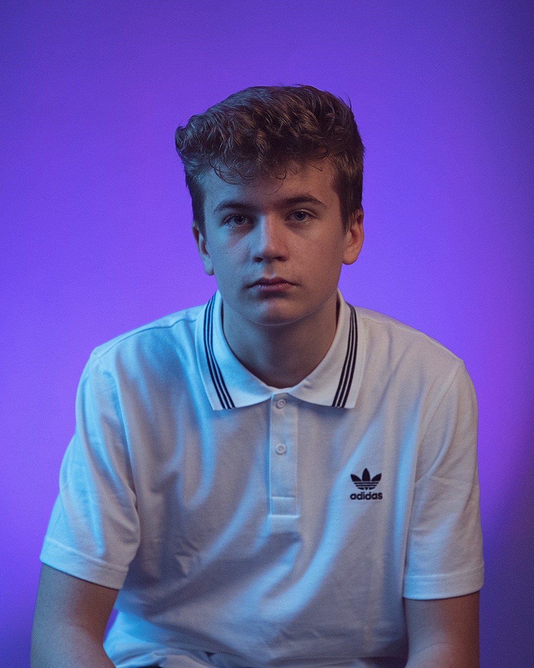 Portret młodego mężczyzny w białej koszulce polo z logo Adidas, na fioletowym tle z subtelnym oświetleniem.