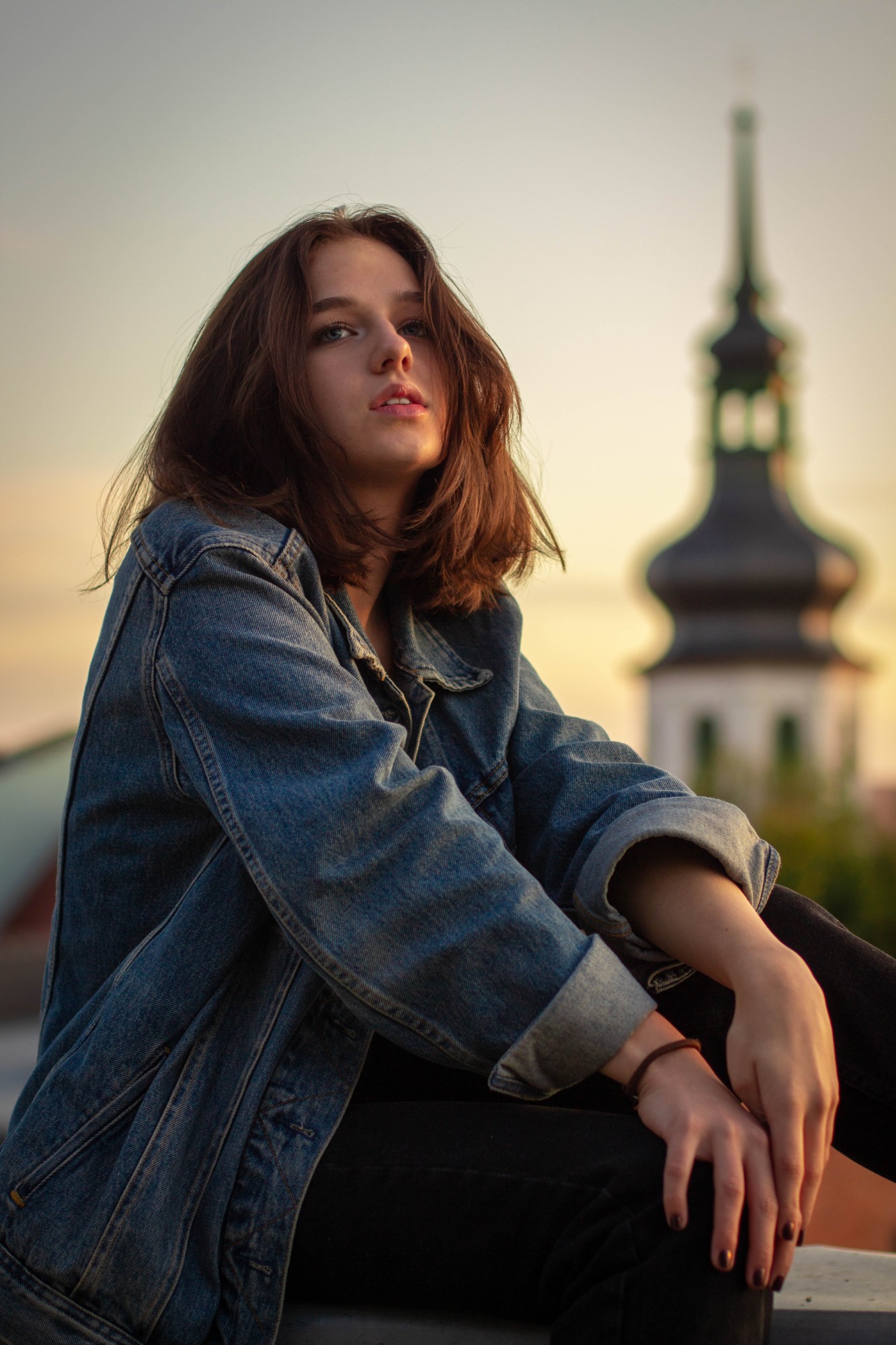 Portret młodej kobiety w dżinsowej kurtce, siedzącej na tle rozmytej wieży kościelnej o zachodzie słońca.