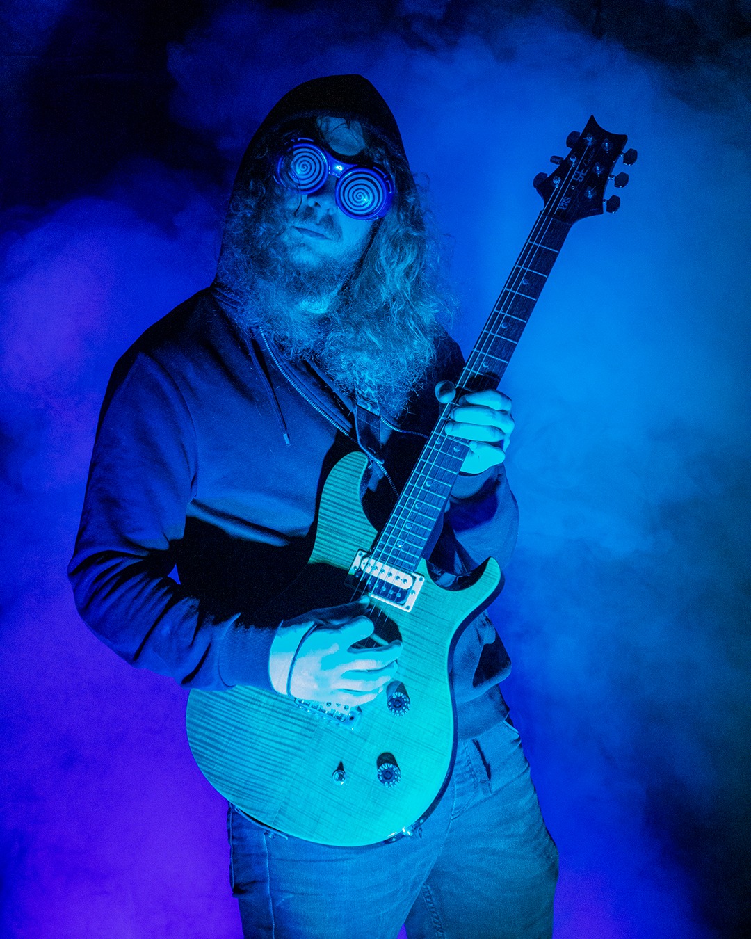 Mężczyzna z długimi włosami i brodą w kapturze i spiralnych okularach gra na elektrycznej gitarze w niebieskim oświetleniu i dymie.