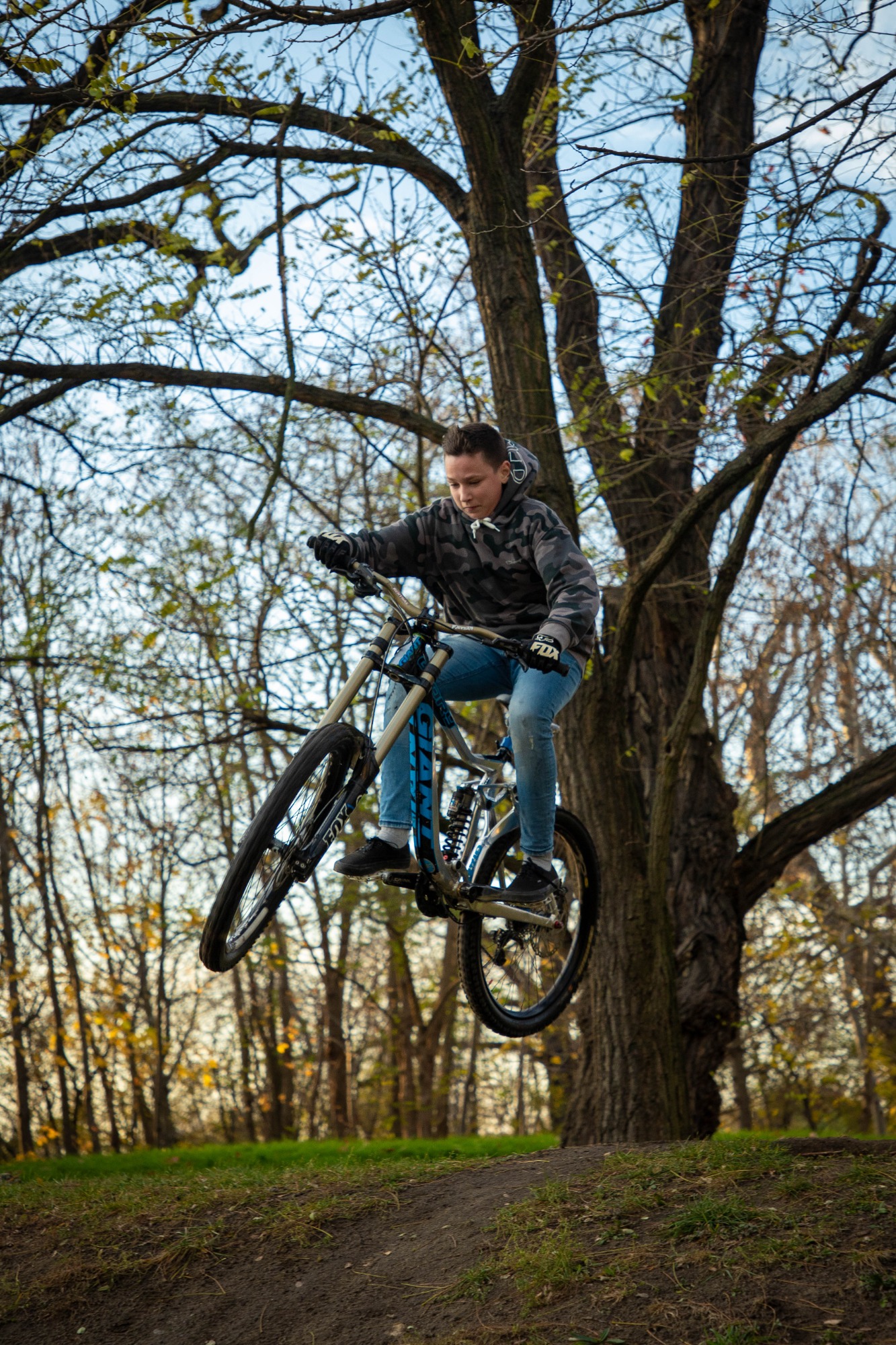 Młody rowerzysta w kamuflażowej bluzie wykonuje skok na rowerze górskim na tle drzew w parku.
