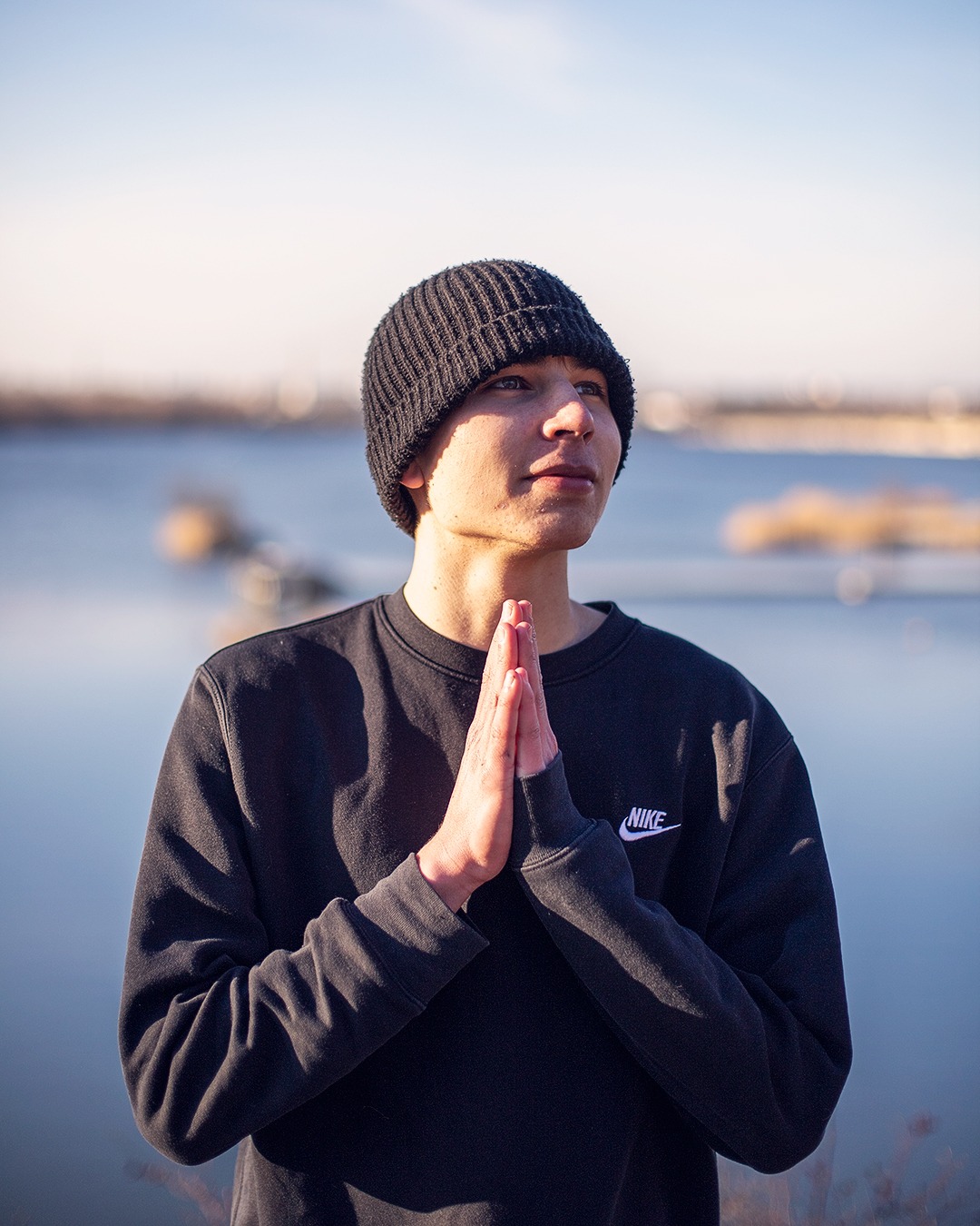 Młody mężczyzna w czarnej czapce i bluzie Nike, z dłońmi złożonymi w geście modlitewnym, spogląda w górę na tle jeziora i błękitnego nieba.