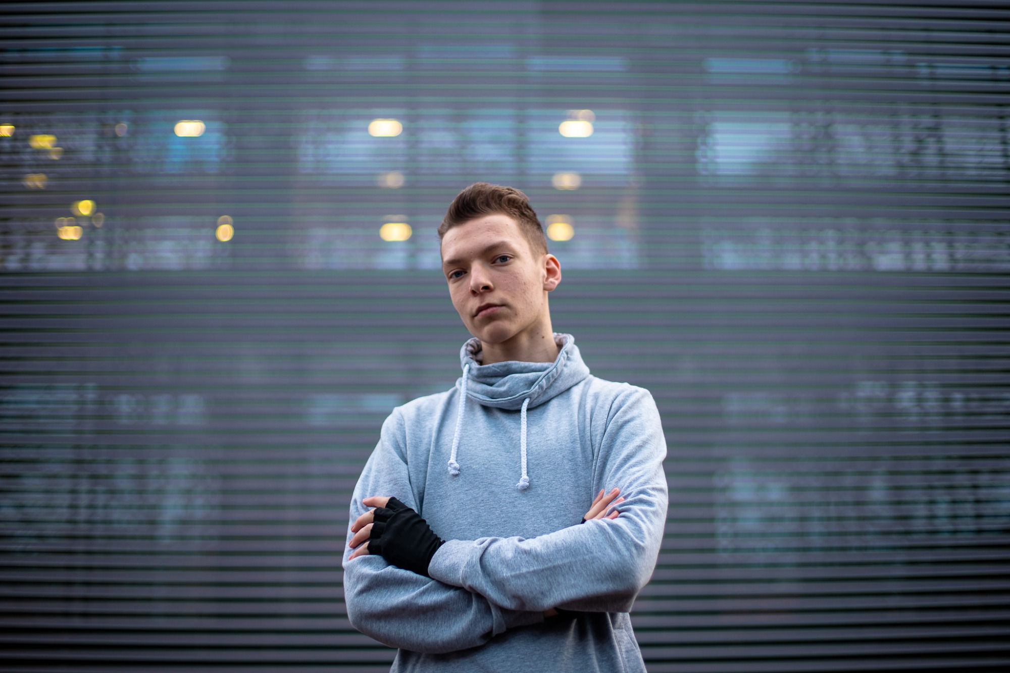 Portret młodego mężczyzny w szarej bluzie z golfem, skrzyżowane ręce w czarnych rękawiczkach bez palców, na tle elewacji budynku z poziomymi liniami i rozmytymi światłami.