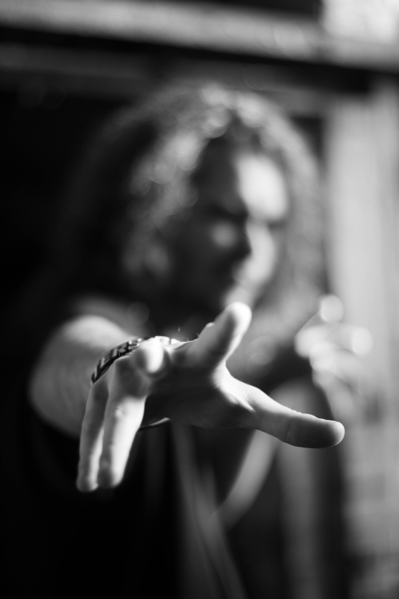 Czarno-białe ujęcie dłoni wyciągniętej do przodu, z rozmytą postacią w tle i bransoletką na nadgarstku.
