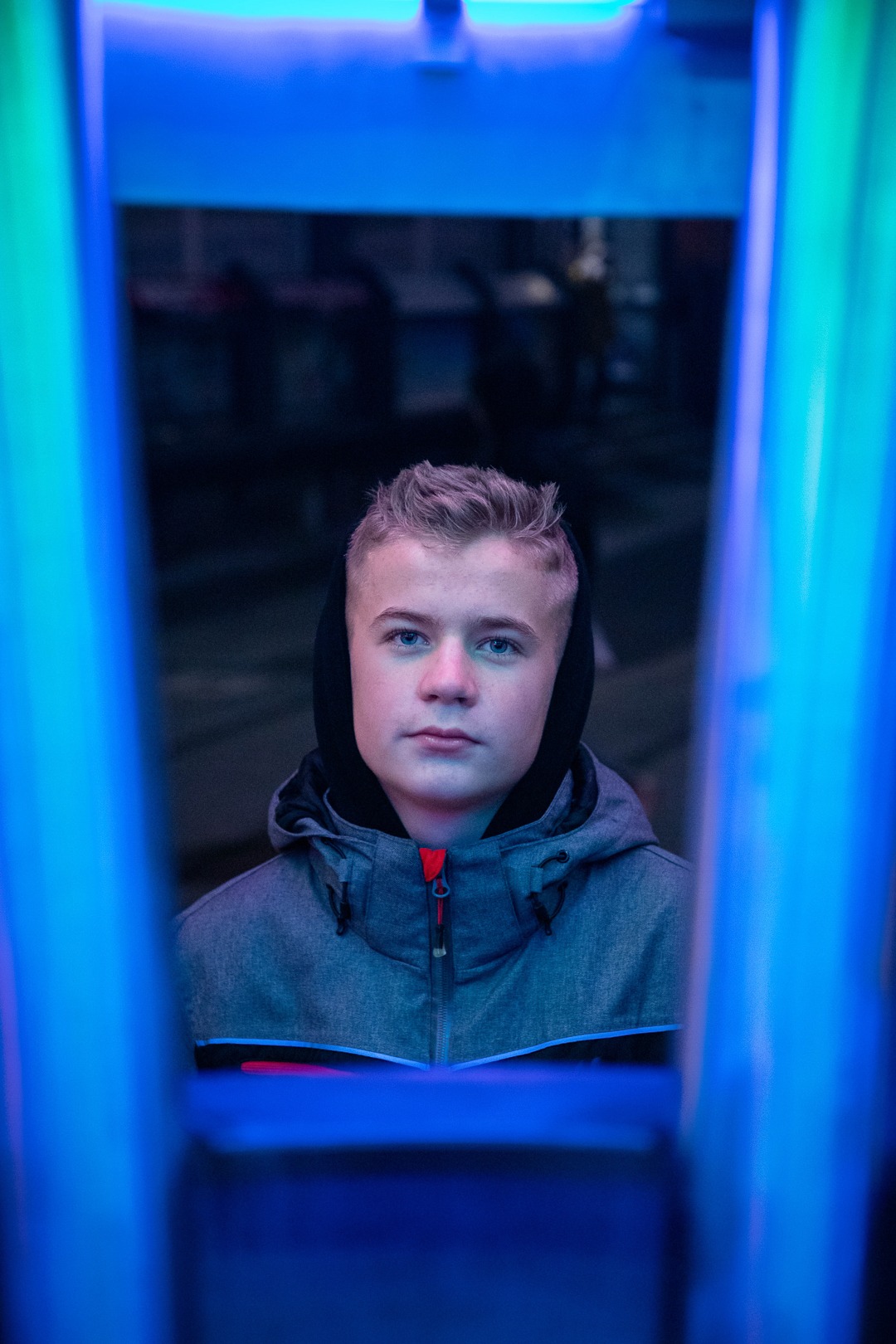 Portret młodego mężczyzny w szarej kurtce z kapturem, oświetlonego niebieskim neonem, z poważnym wyrazem twarzy.