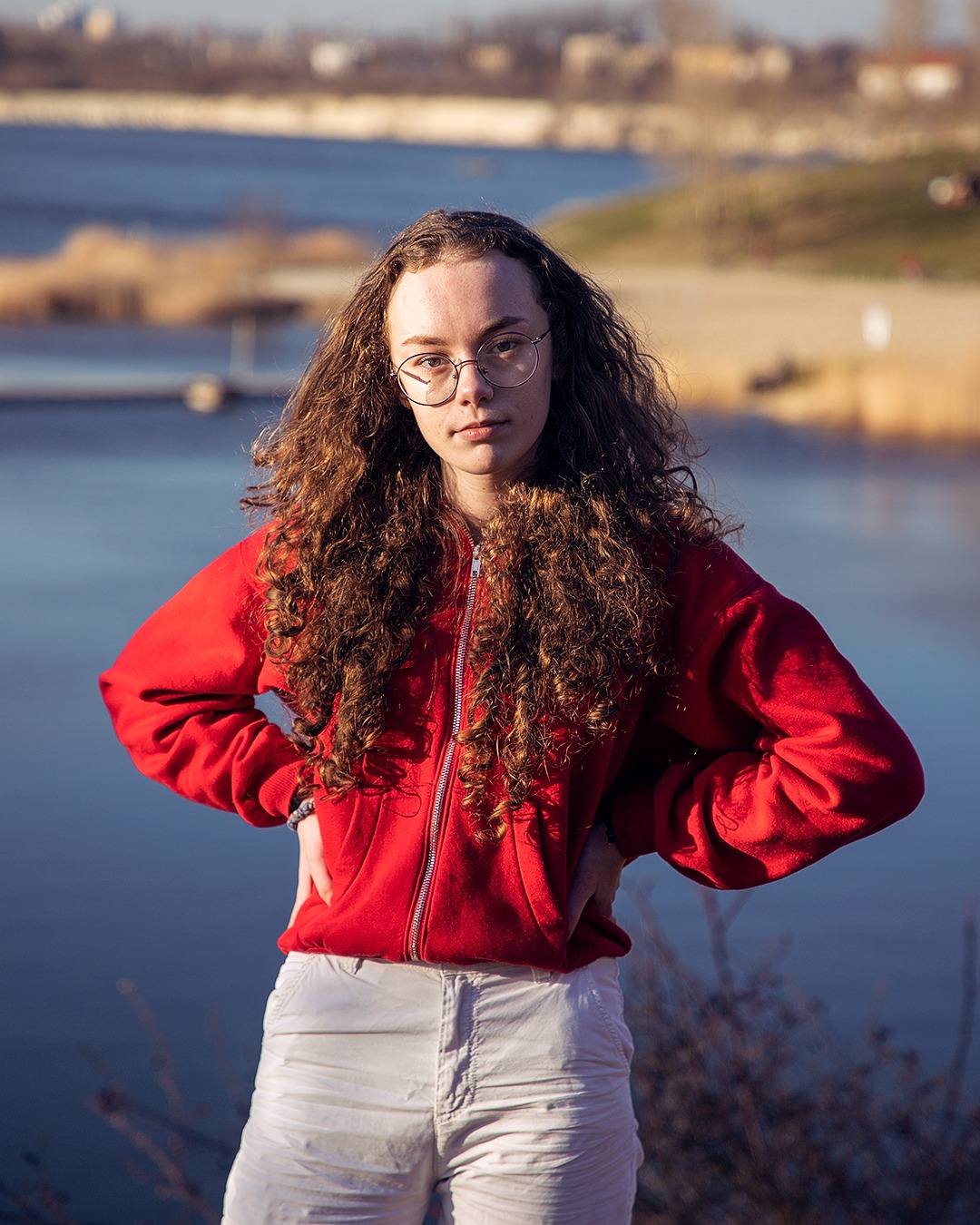 Portret młodej kobiety w okularach, ubrana w czerwoną bluzę i białe spodnie, pozująca na tle jeziora i oddalonego krajobrazu miejskiego.