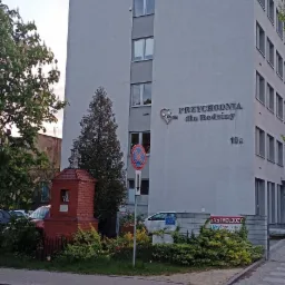 Szary budynek przychodni z napisem 'Przychodnia dla Rodziny 19A', obok czerwona kapliczka, znak zakazu zatrzymywania się i tablica informacyjna o gastrologu.