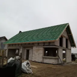 Niedokończony budynek jednorodzinny z białego bloczka, z częściowo ułożonym zielonym pokryciem dachowym i pracownikiem na dachu, widok z zewnątrz w pochmurny dzień.