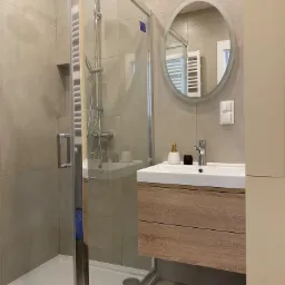 Nowoczesna łazienka w beżowych odcieniach z przeszkloną kabiną prysznicową, umywalką z szafką z drewnianym dekorem, okrągłym lustrem z podświetleniem i minimalistyczną armaturą.