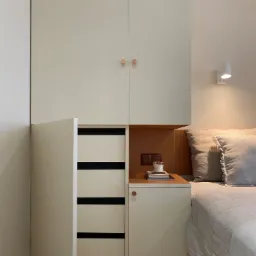 Zabudowa szafy wnękowej z szafką nocną w neutralnych barwach, z elementami drewna i minimalistycznym oświetleniem, uzupełniona o świecę i książki na blacie.