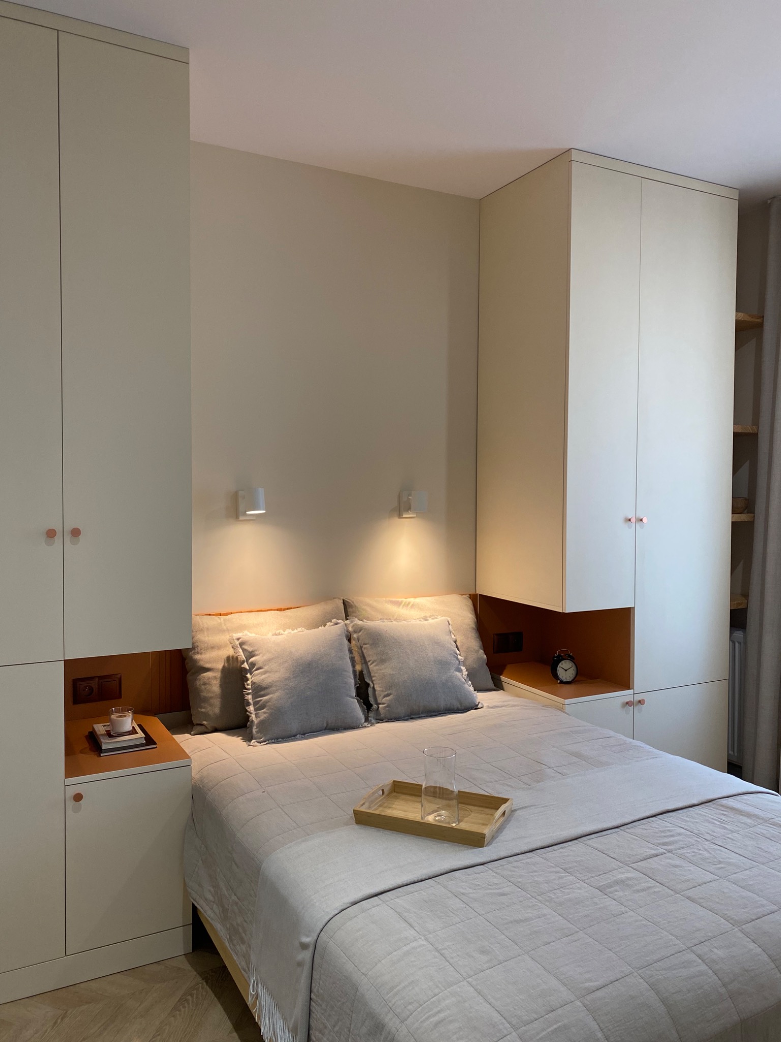 Nowoczesna sypialnia z zabudową meblową w odcieniach beżu, obejmująca szafy po bokach łóżka, szafki nocne z drewnianymi akcentami i dekoracyjnym oświetleniem ściennym.