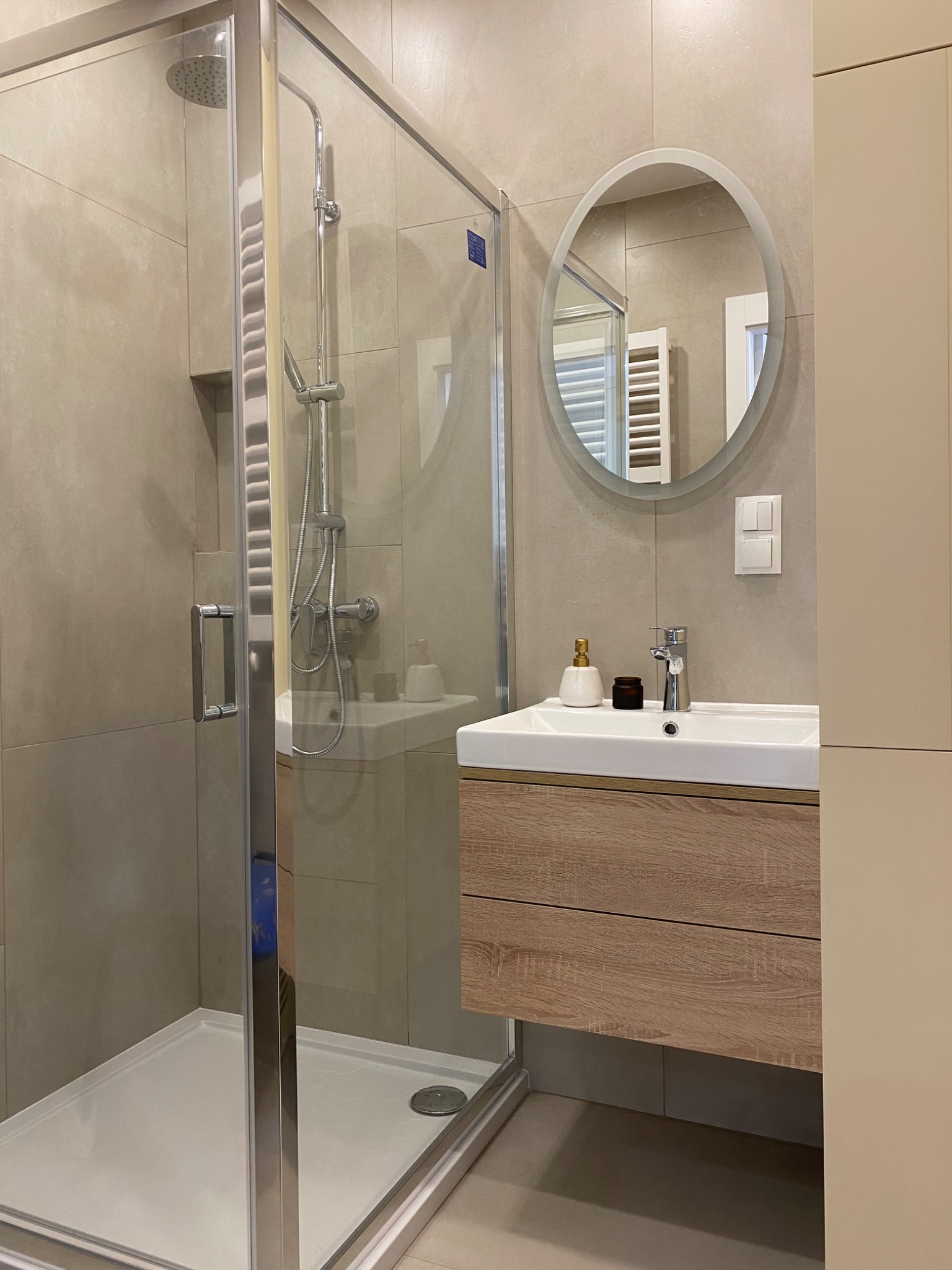 Nowoczesna łazienka w beżowych odcieniach z przeszkloną kabiną prysznicową, umywalką z szafką z drewnianym dekorem, okrągłym lustrem z podświetleniem i minimalistyczną armaturą.