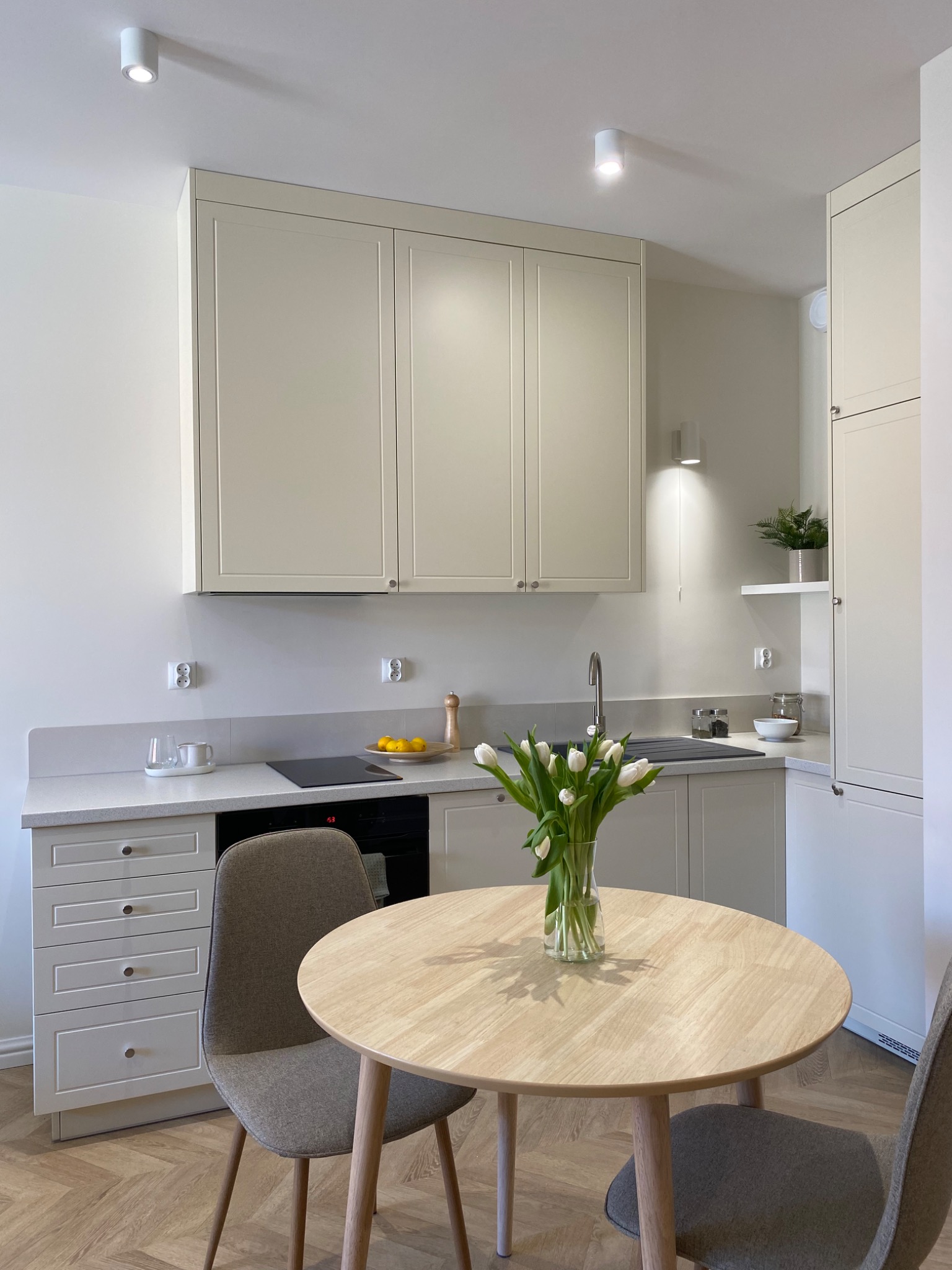 Jasna kuchnia z drewnianym okrągłym stołem i bukietem białych tulipanów, minimalistyczny design, parkiet ułożony w jodełkę.