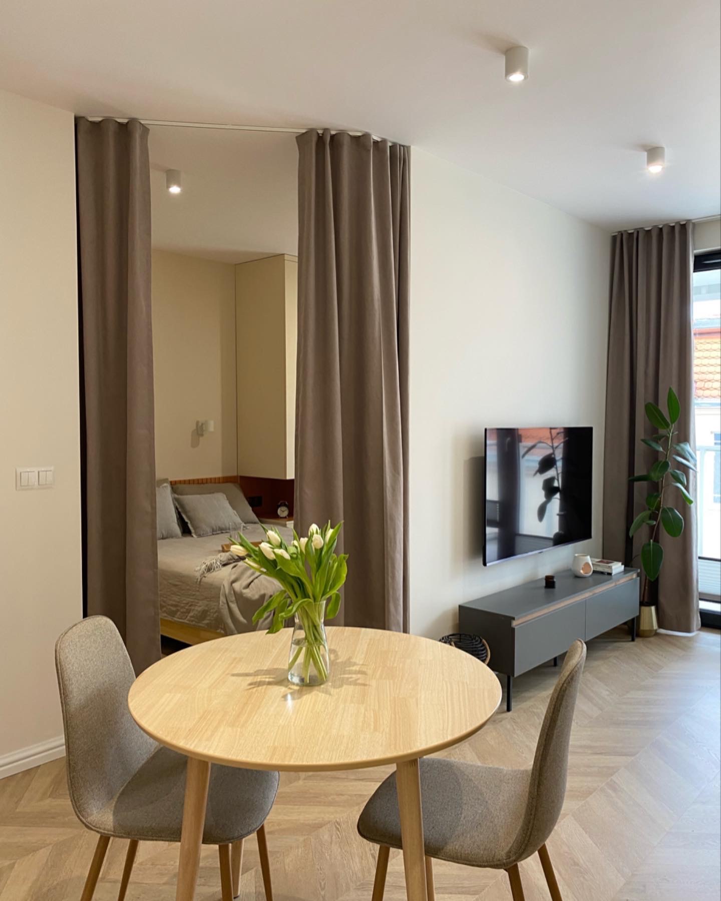 Jasne wnętrze mieszkania z wydzieloną strefą sypialnianą za pomocą zasłon, okrągły stół z bukietem białych tulipanów na pierwszym planie, podłoga ułożona w jodełkę.