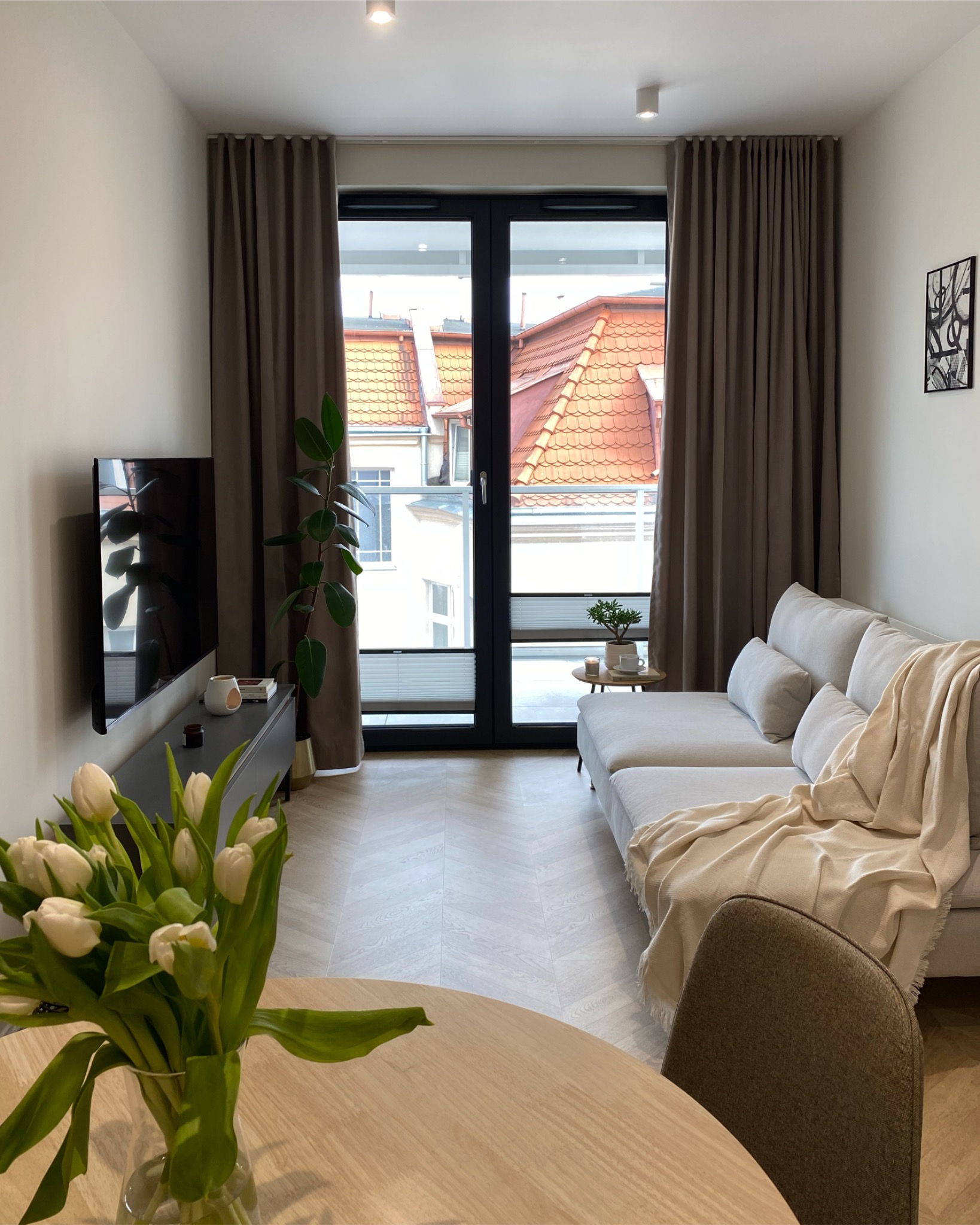 Jasne, nowocześnie urządzone mieszkanie z drewnianą podłogą w jodełkę, szarą sofą z beżowym pledem, okrągłym stołem z bukietem białych tulipanów i widokiem na czerwone dachy sąsiednich budynków.