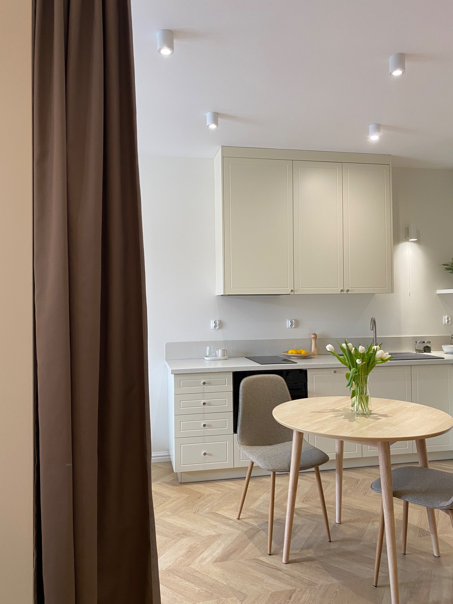 Jasna kuchnia z drewnianym stołem i bukietem białych tulipanów, widok częściowo zasłonięty brązową zasłoną, parkiet w jodełkę.