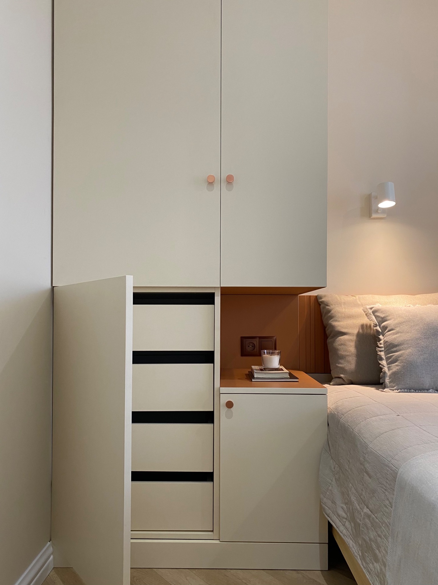 Zabudowa szafy wnękowej z szafką nocną w neutralnych barwach, z elementami drewna i minimalistycznym oświetleniem, uzupełniona o świecę i książki na blacie.