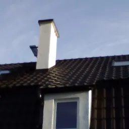 Ciemny dach z dachówki ceramicznej z kominem i oknami dachowymi, widok z dołu na tle jasnego nieba.