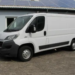 Biały furgon Fiat Ducato zaparkowany przed budynkiem z panelami słonecznymi na dachu, widoczny z boku na zewnątrz.