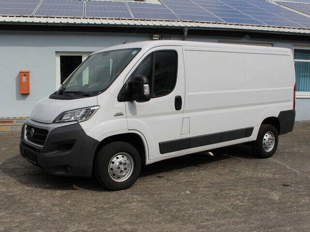 Biały furgon Fiat Ducato zaparkowany przed budynkiem z panelami słonecznymi na dachu, widoczny z boku na zewnątrz.