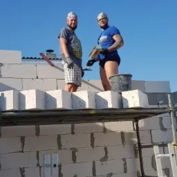 Dwóch mężczyzn na rusztowaniu, murują ścianę z białych bloczków betonowych, widoczna drabina i niebieskie niebo w tle.