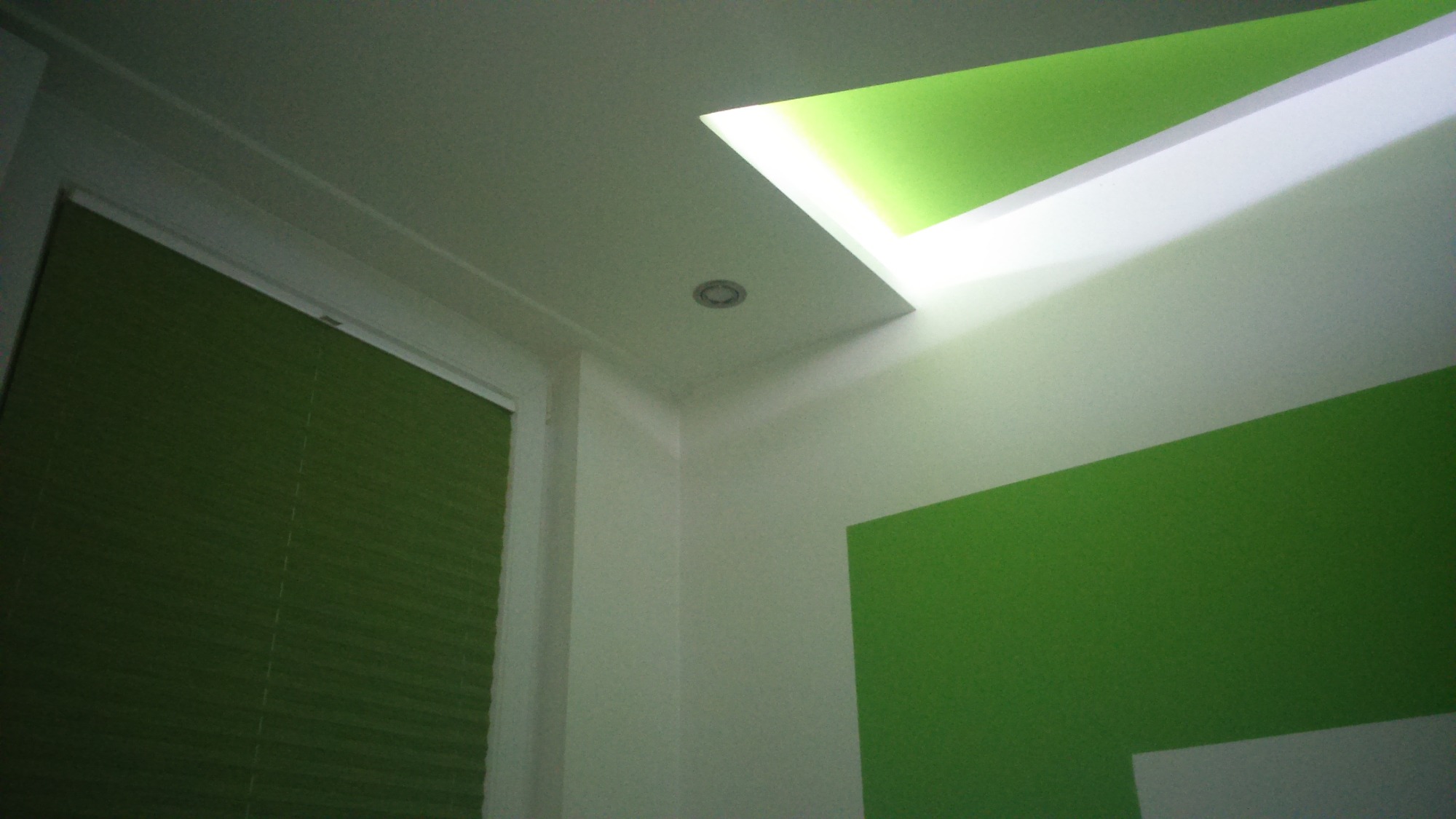 Fragment wnętrza z zielonymi akcentami: pomalowana ściana, roleta w oknie i podświetlany fragment sufitu.