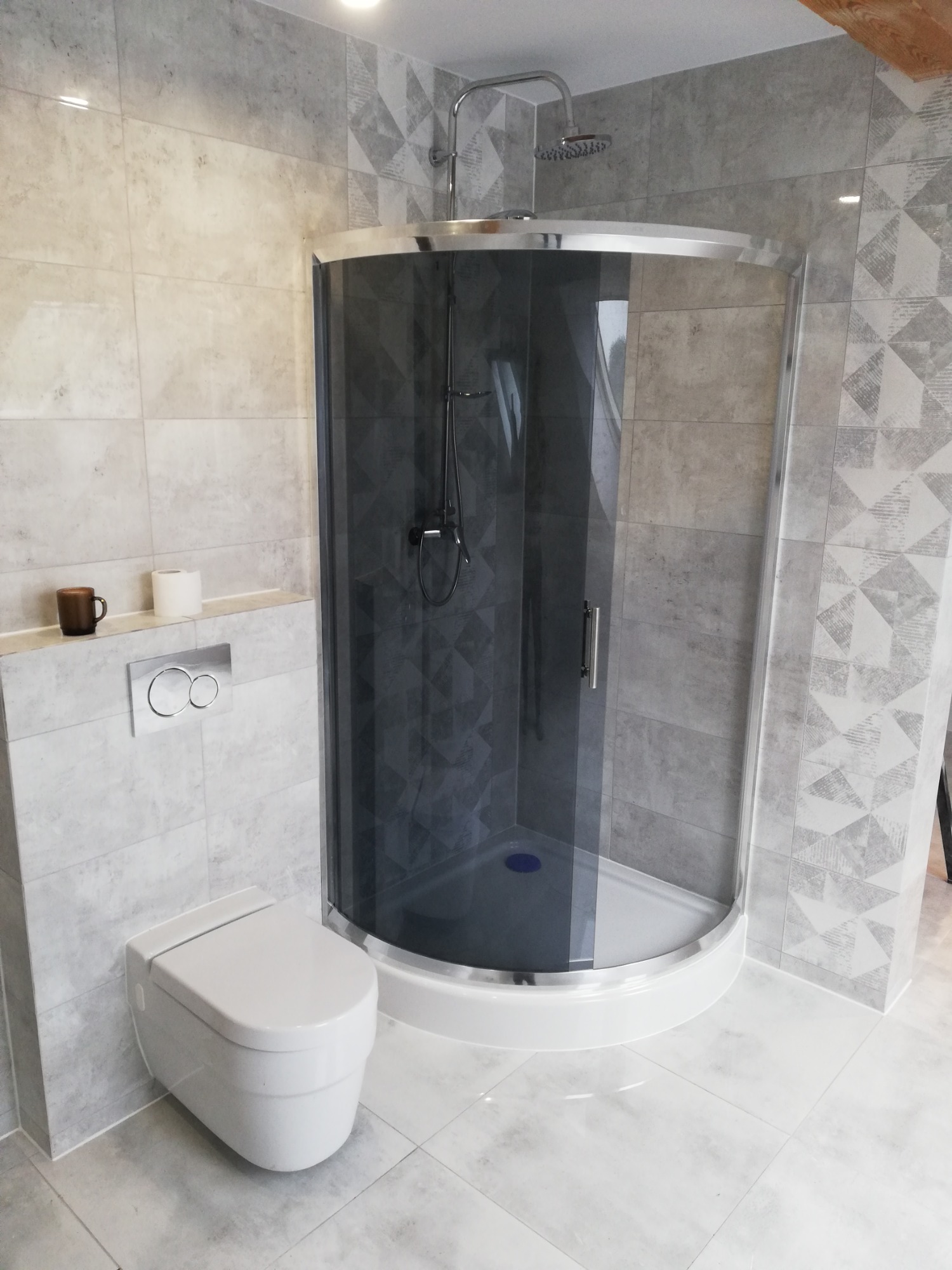 Nowoczesna łazienka z kabiną prysznicową typu walk-in z ciemnymi szybami, wiszącą toaletą i płytkami w odcieniach szarości z geometrycznymi wzorami.