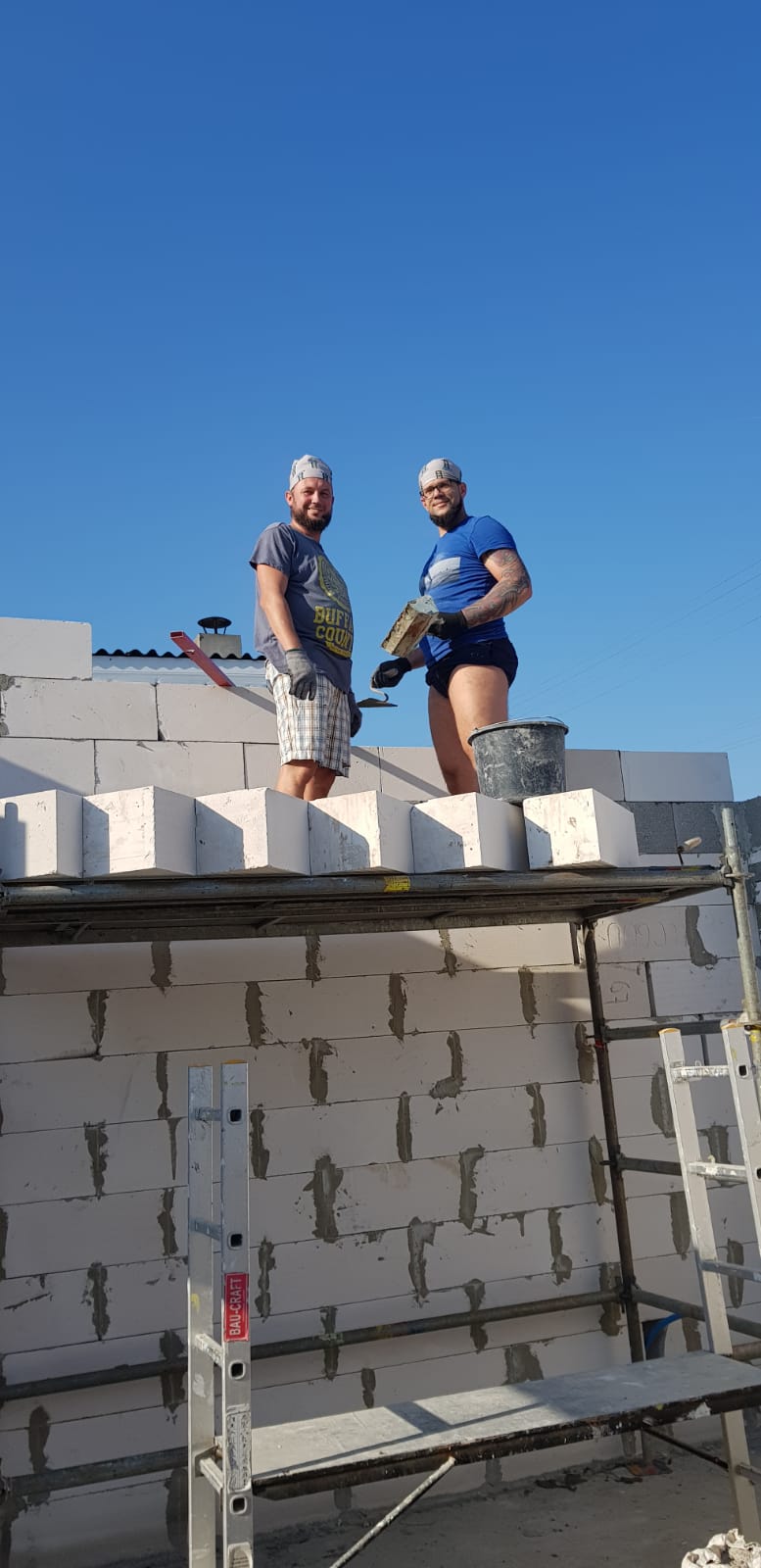 Dwóch mężczyzn na rusztowaniu, murują ścianę z białych bloczków betonowych, widoczna drabina i niebieskie niebo w tle.