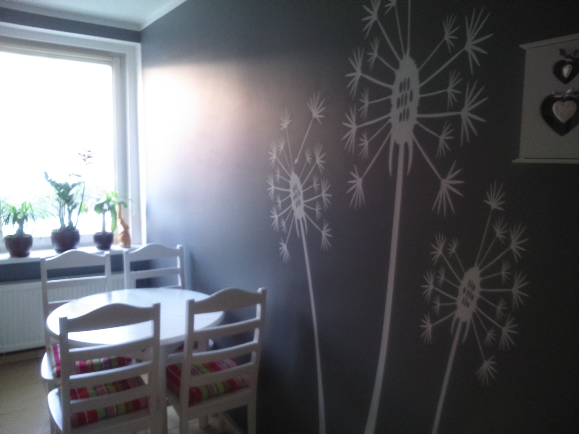 Szary pokój z pomalowaną ścianą w białe dmuchawce, biały okrągły stół z krzesłami i kolorowymi poduszkami, oraz półka z dekoracjami w kształcie serc.
