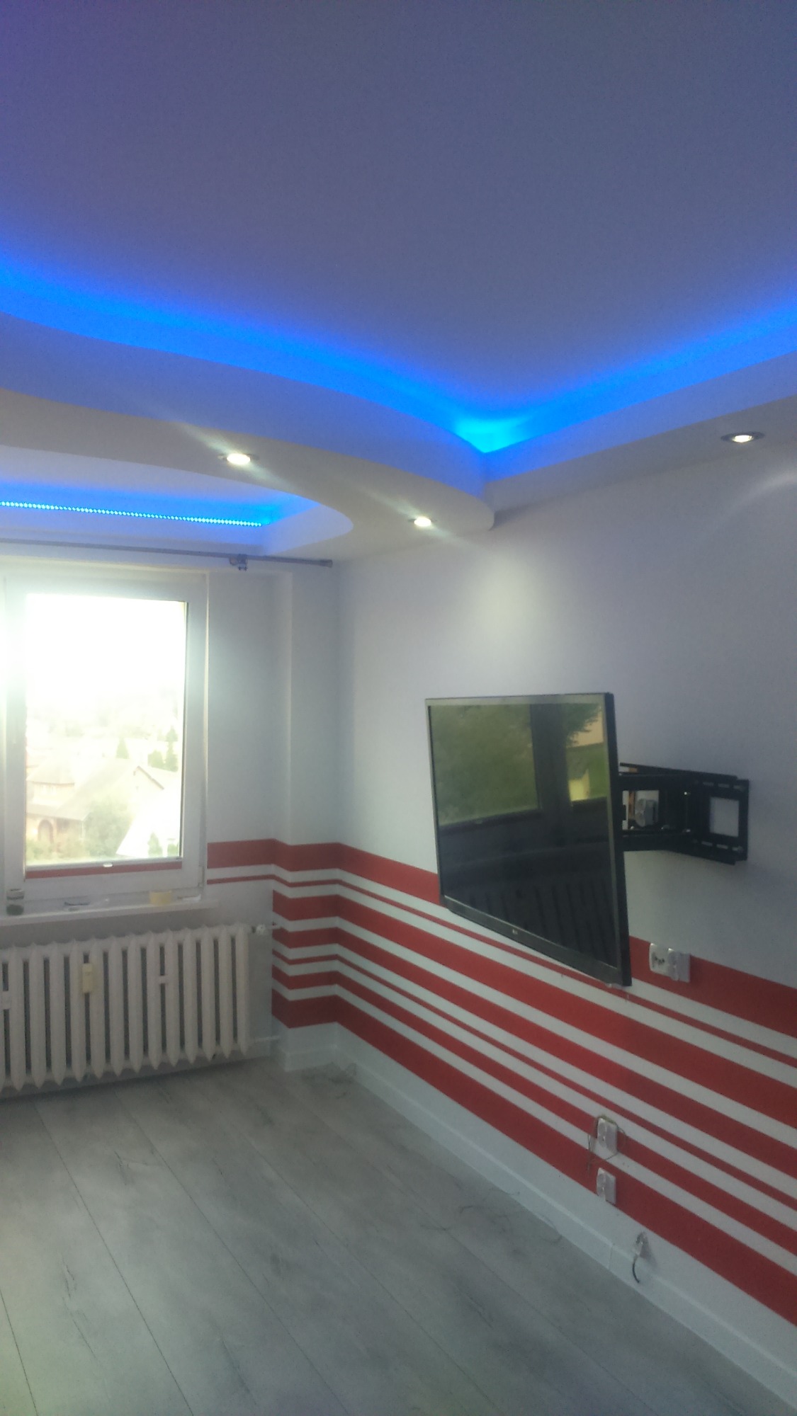 Pokój po remoncie z białymi ścianami ozdobionymi poziomymi czerwonymi pasami, szarymi panelami podłogowymi, grzejnikiem pod oknem i telewizorem wiszącym na ścianie, podwieszany sufit z niebieskim...