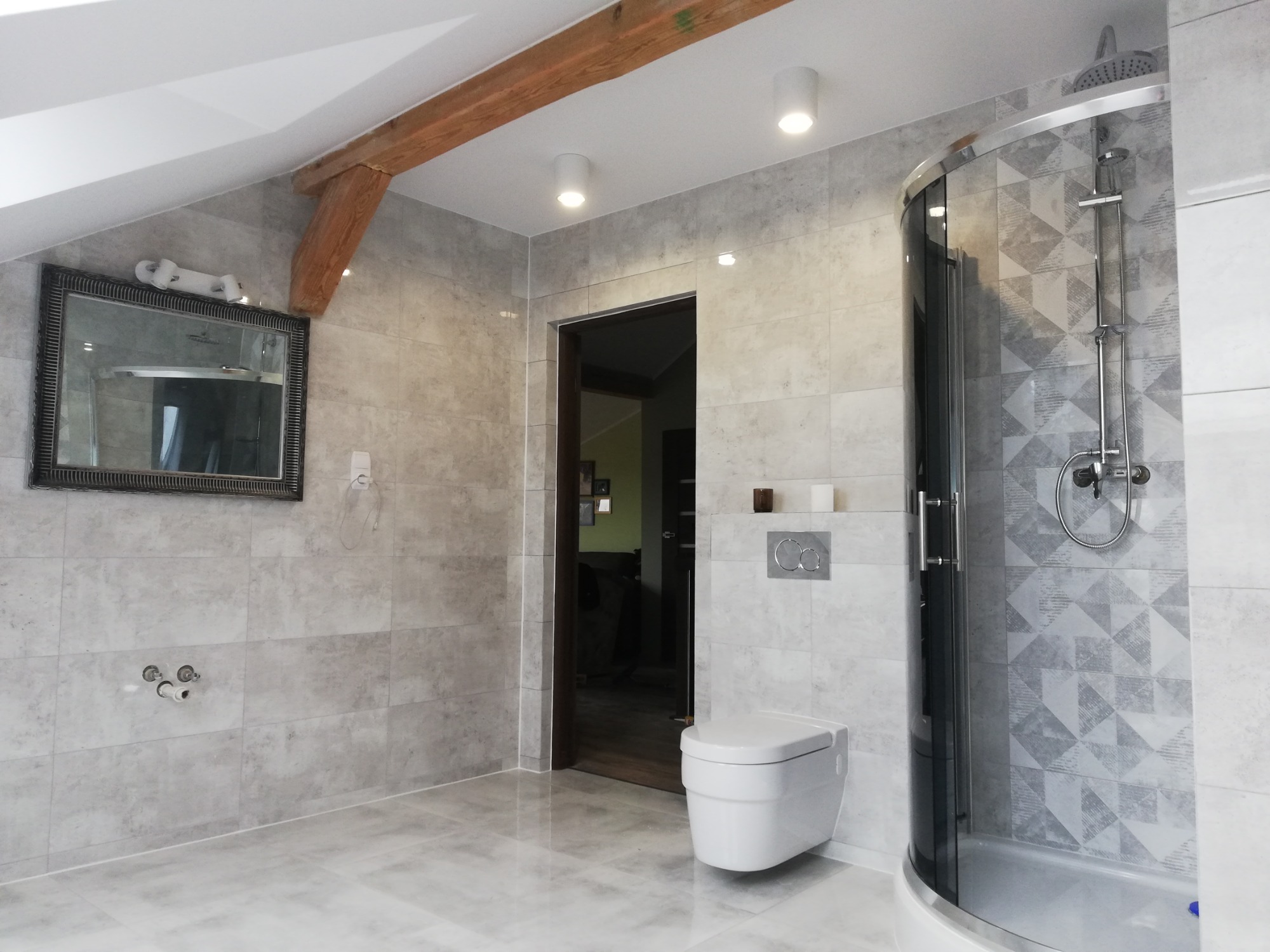 Nowoczesna łazienka na poddaszu z szarą glazurą, kabiną prysznicową z ciemnymi profilami, podwieszaną toaletą i drewnianą belką stropową.