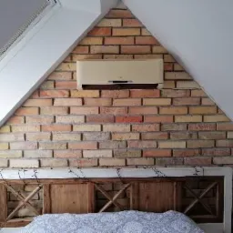 Klimatyzator zamontowany nad wezgłowiem łóżka na ścianie z cegły w sypialni na poddaszu.
